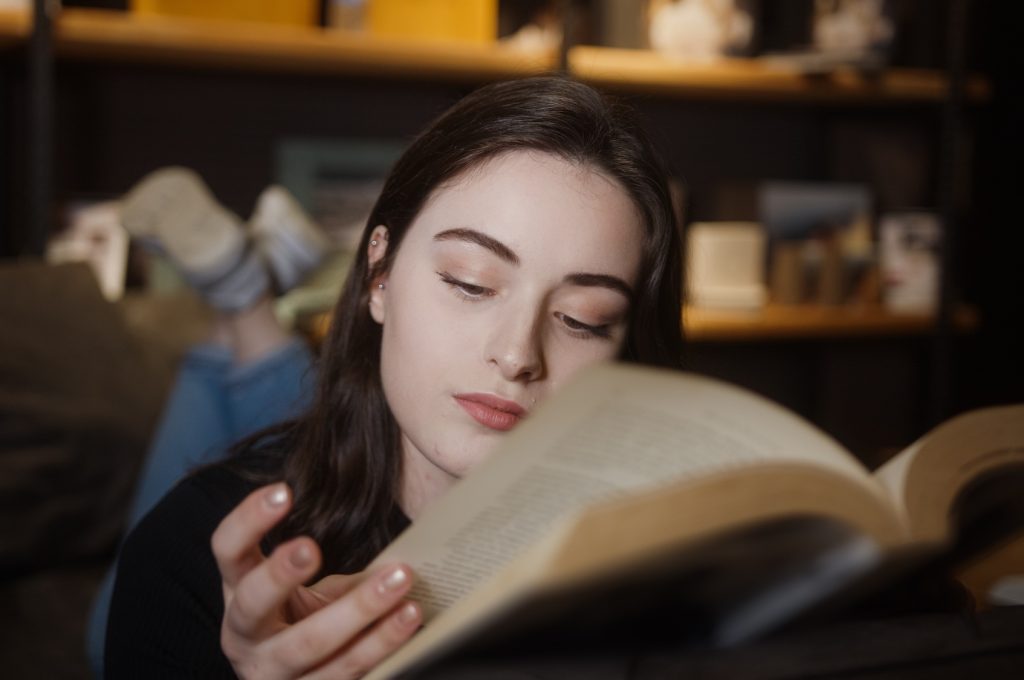 Sweet young woman lying on sofa at home, reading novel or educational book